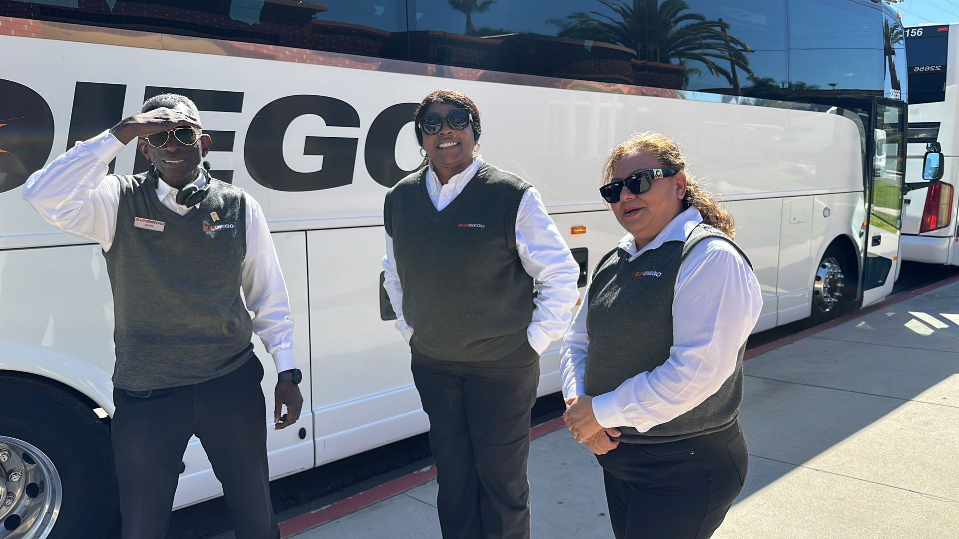 Three people posing beside a white tour bus in bright sunlight, one waving, one smiling.
