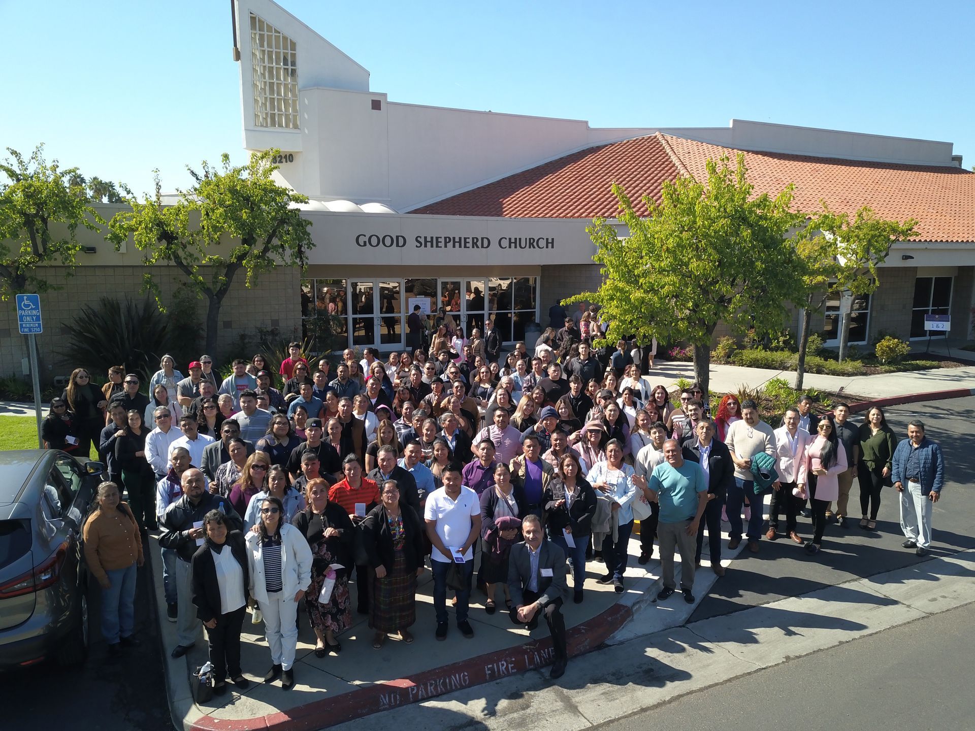 Large group posing outside a building with a red-tiled roof and sign, under a sunny sky