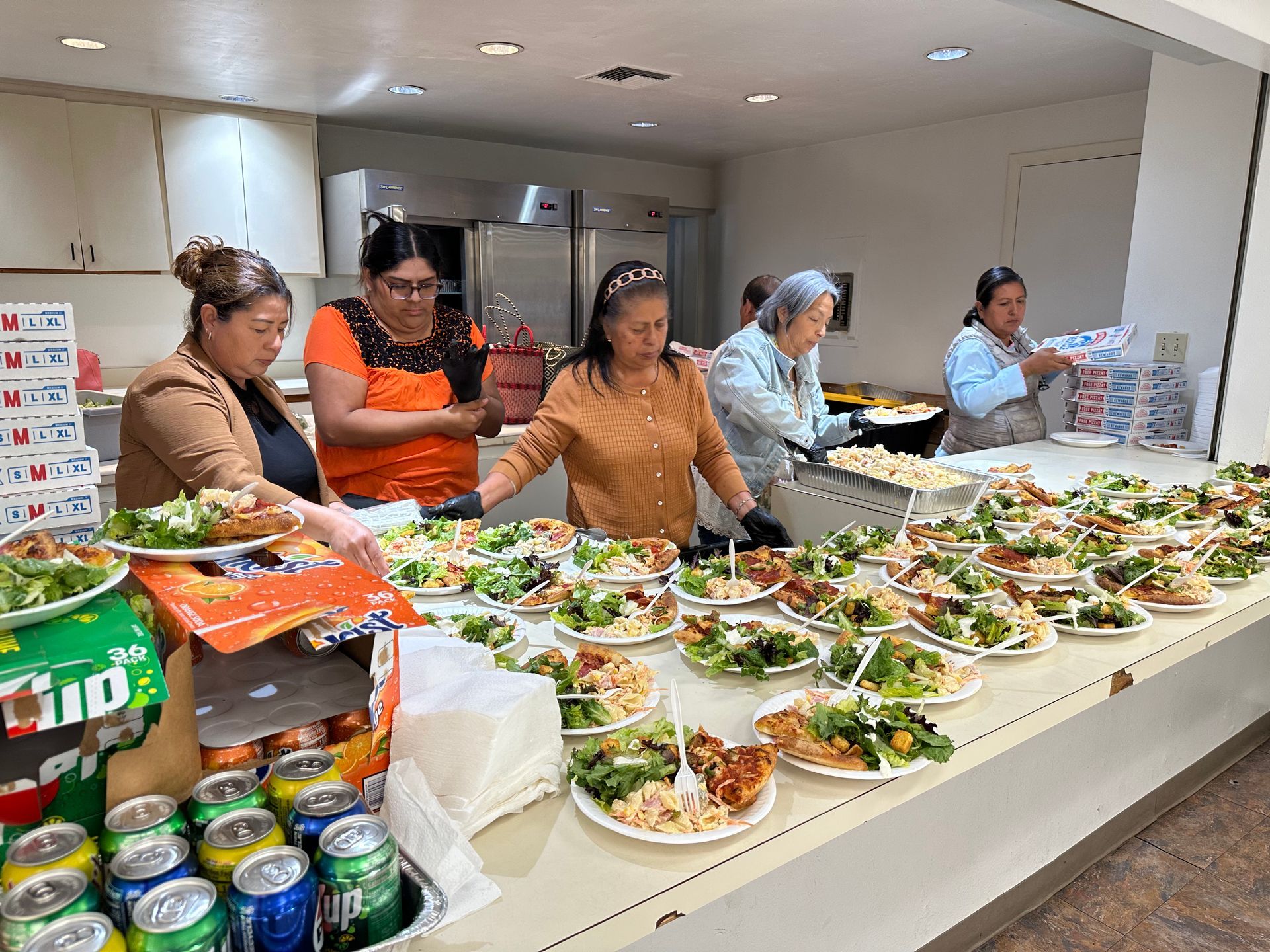 People serving plated food at a buffet table in a bright kitchen, with salads and drinks spread across the counter