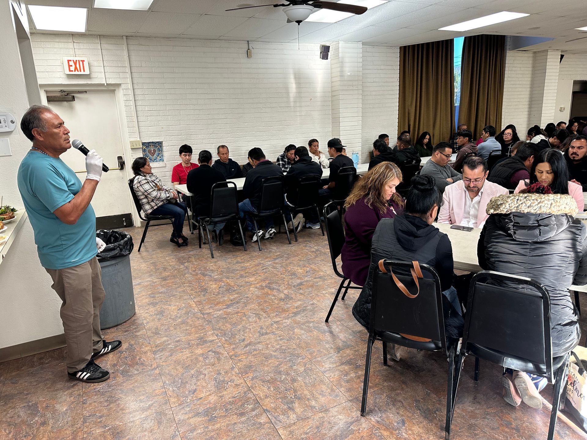 Man speaking to a seated group in a community meeting room with round tables and white brick walls