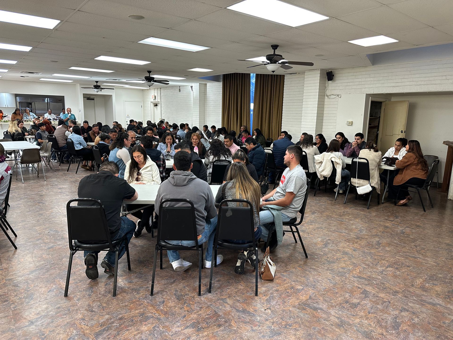 Large community meeting in a banquet hall, with seated attendees around circular tables under bright fluorescent lights.