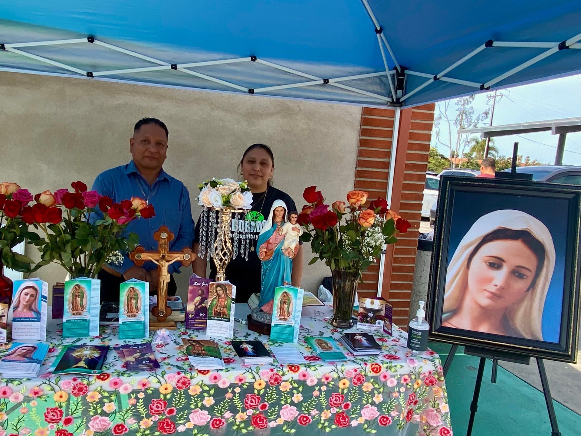 Outdoor memorial altar under blue canopy with flowers, candles, framed portraits, and a large Virgin Mary image