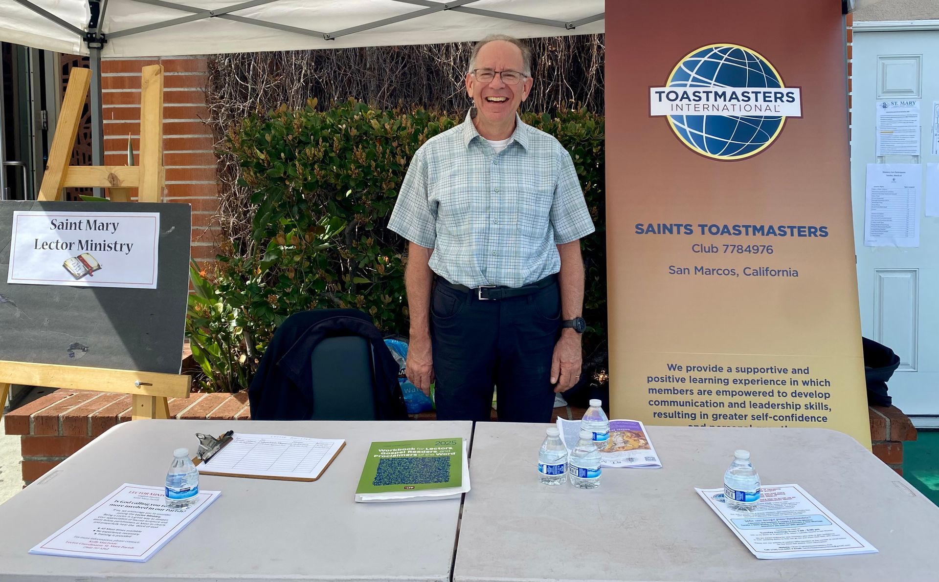 Man smiling behind an expo table with brochures and a blue banner under a white tent.