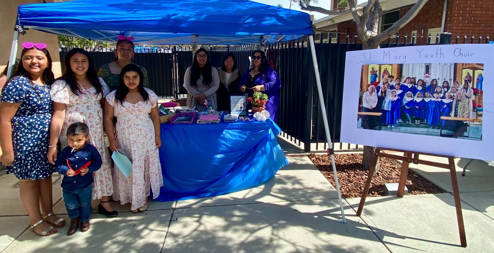 Group posing under blue canopy beside a decorated table and framed photo display outdoors