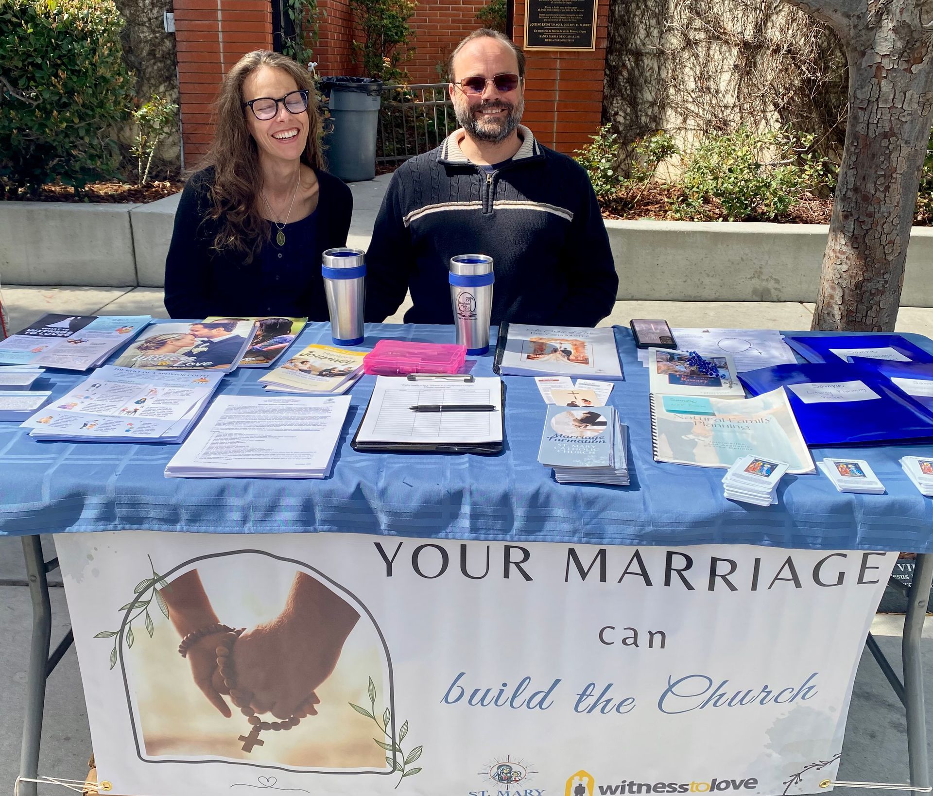 Two people at a church outreach table with pamphlets and cups; banner reads “Your Marriage can Build the Church.”