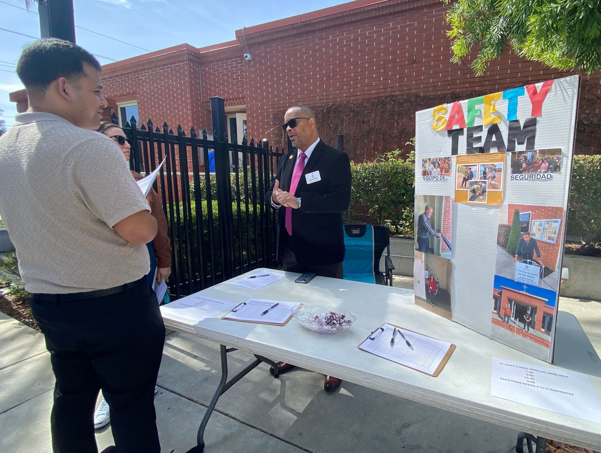 Two people at an outdoor community table beside a colorful “SAFETY TEAM” display board.