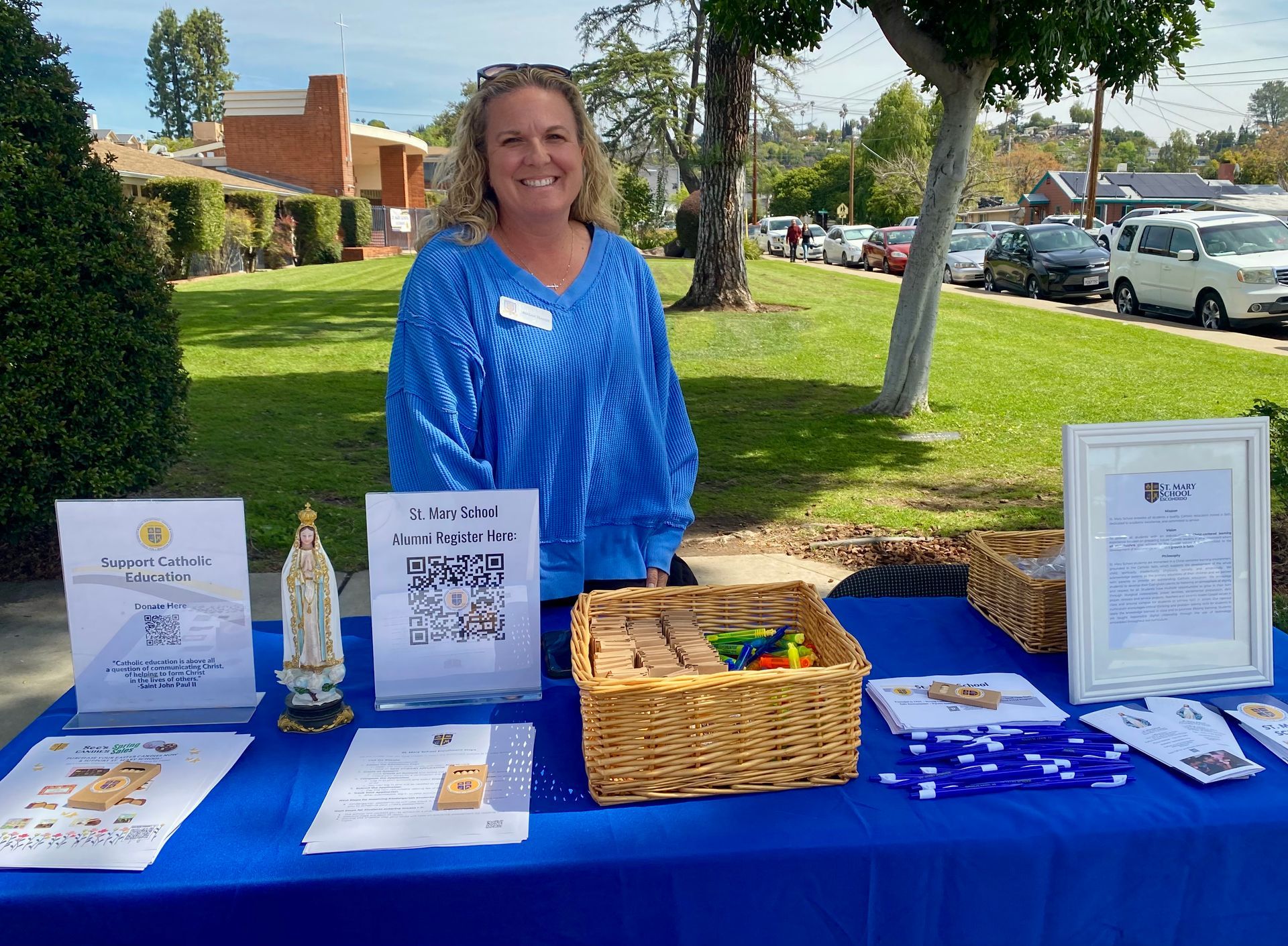 Woman behind blue table at outdoor fair, with flyers, basket, and display items on lawn.