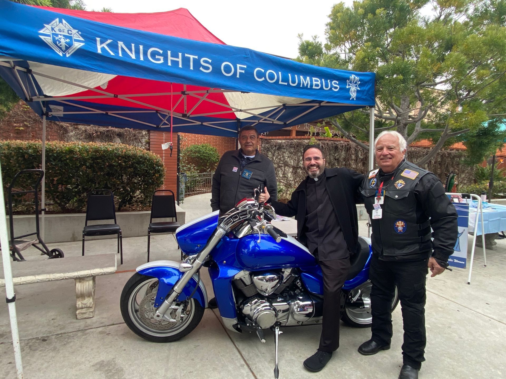 Three men stand beside a blue motorcycle under a Knights of Columbus canopy.