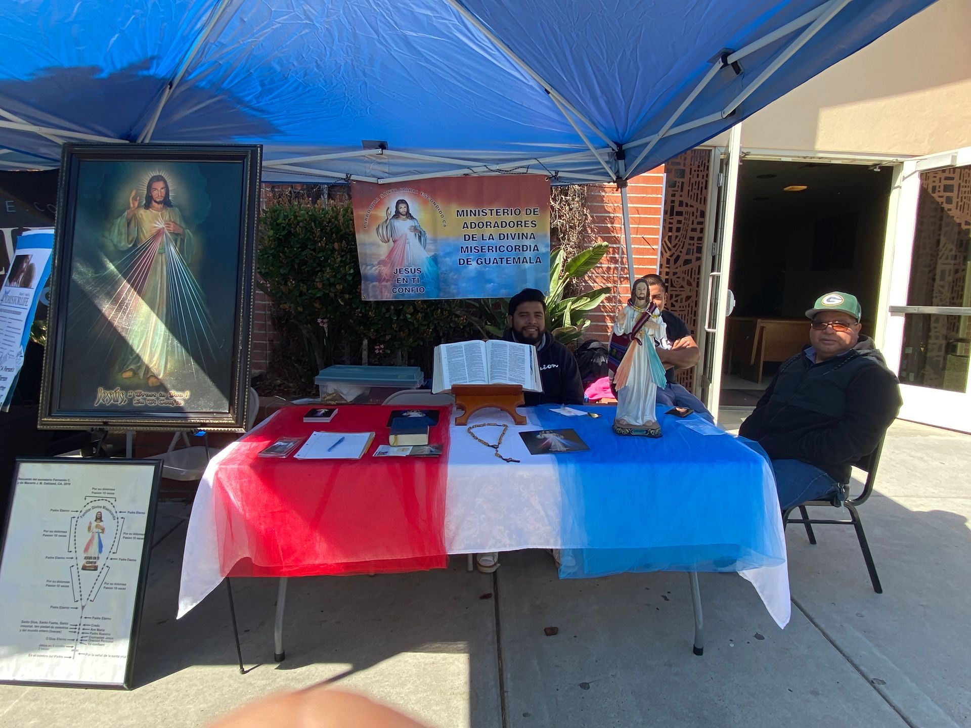 Outdoor booth under blue canopy with religious posters, table displays, and two people seated behind it