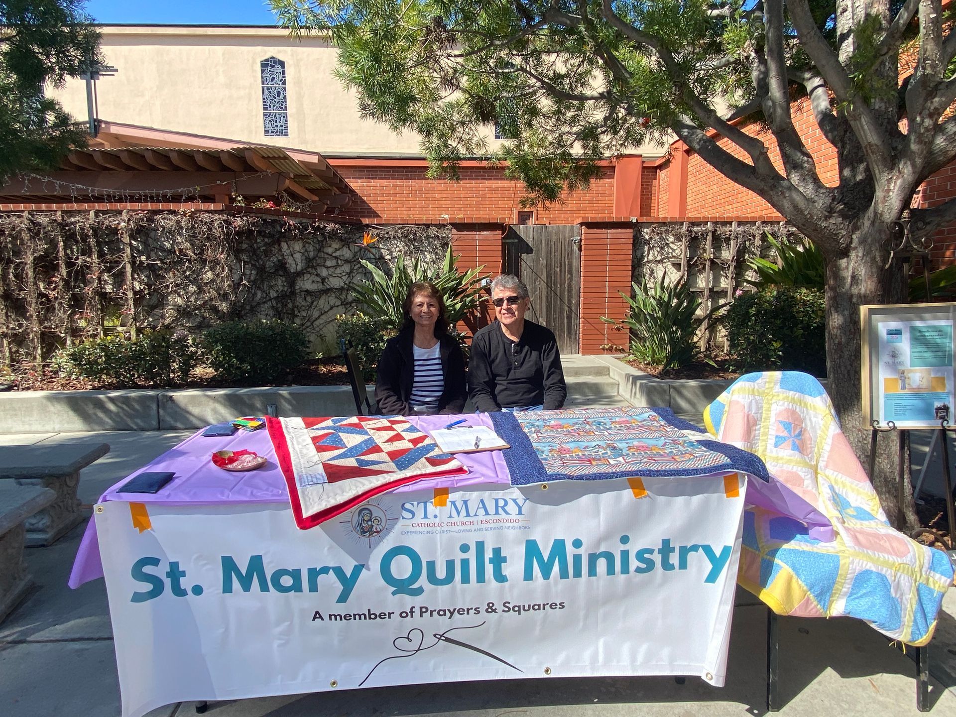 Two people at a table with a “St. Mary Quilt Ministry” banner and colorful quilt display outdoors
