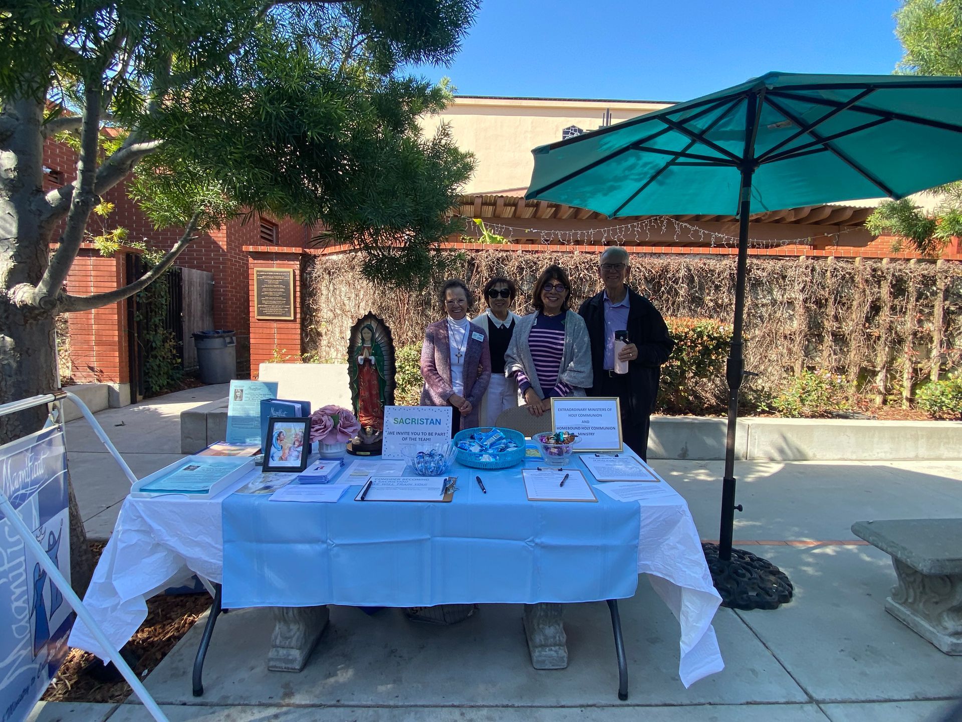 Outdoor booth with four people behind a table under a turquoise umbrella, displaying flyers and books