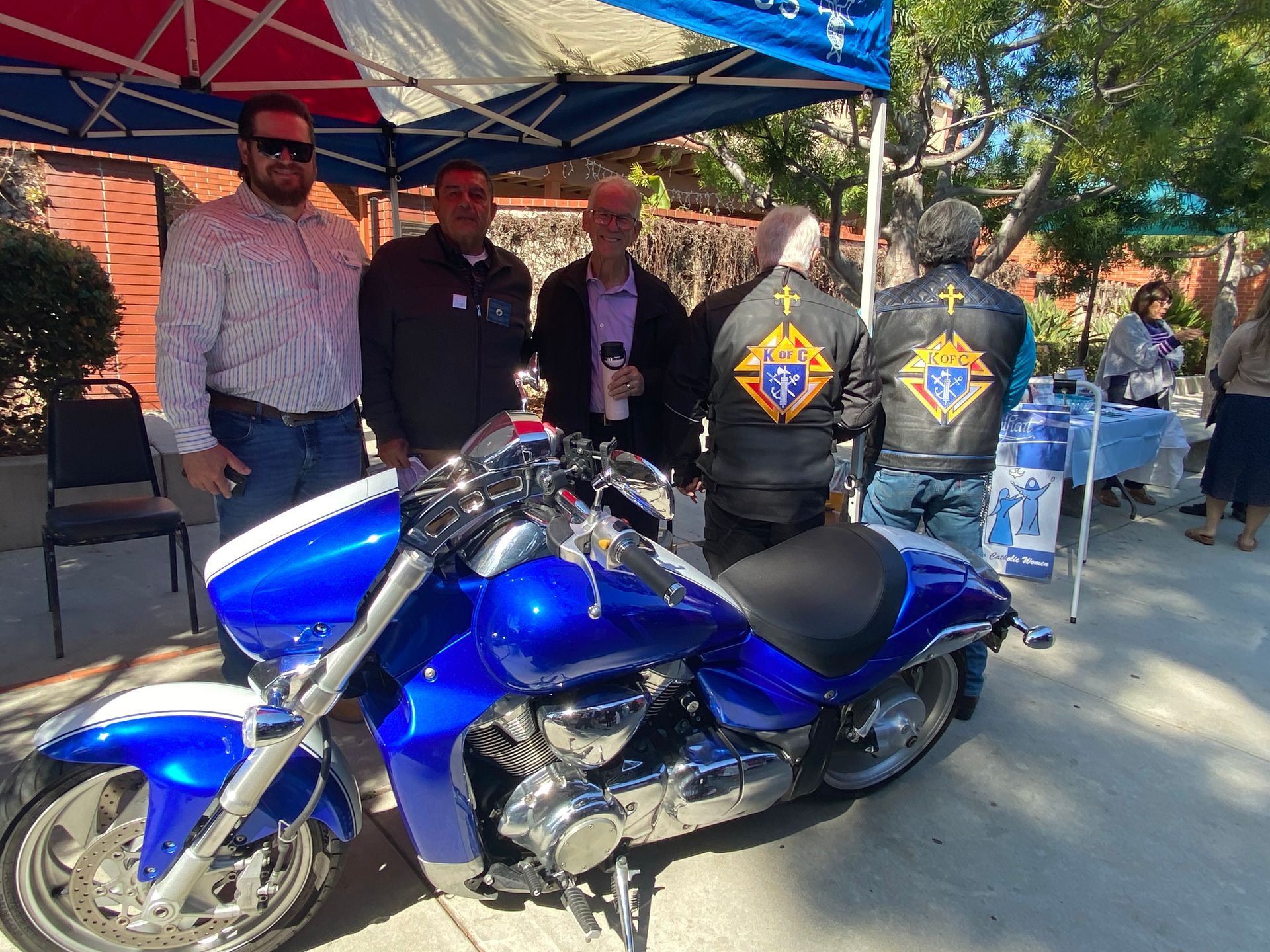 Blue motorcycle displayed with three people standing behind it under a canopy at an outdoor event