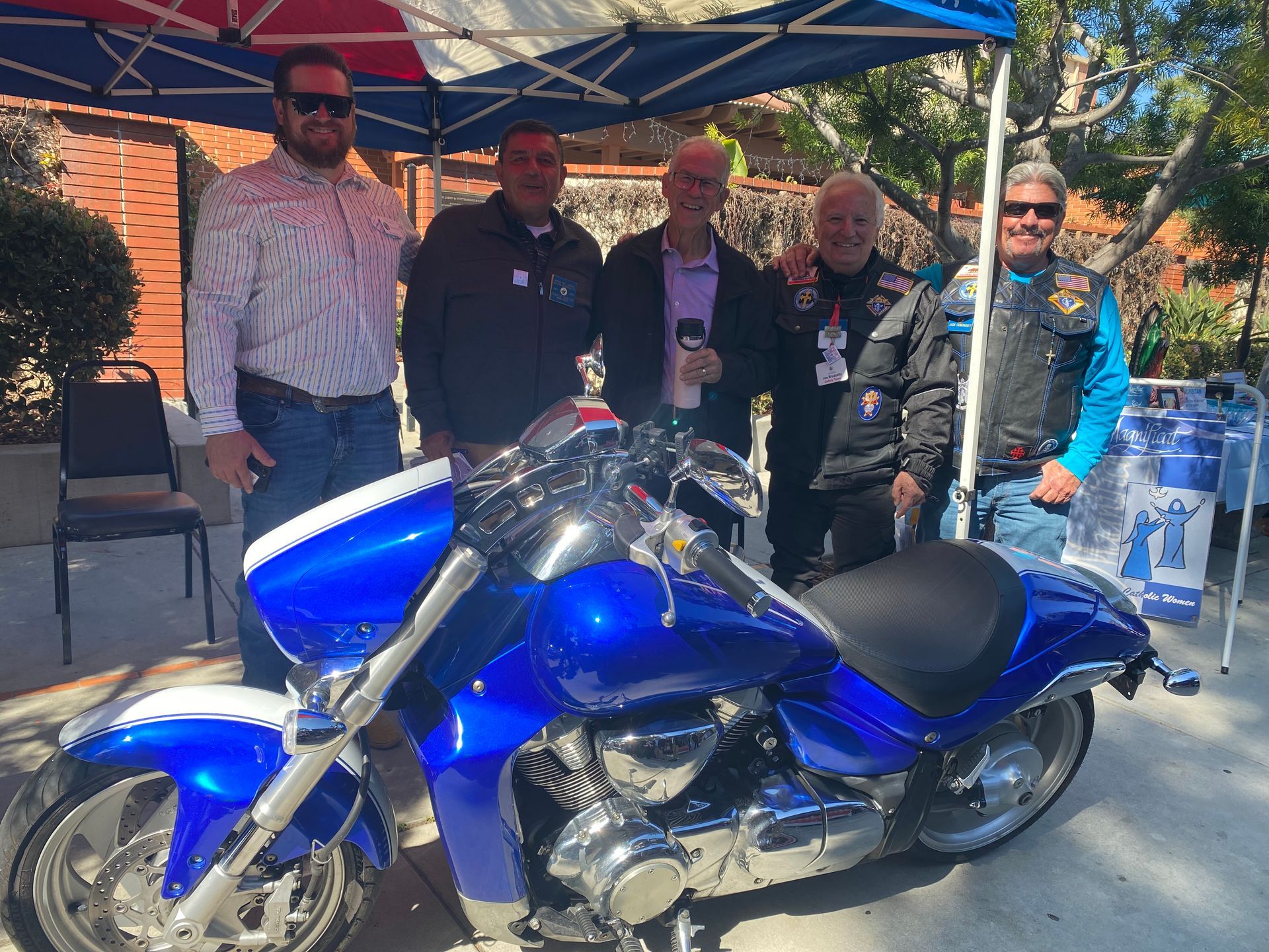 Five people posing behind a blue motorcycle under a canopy at an outdoor event