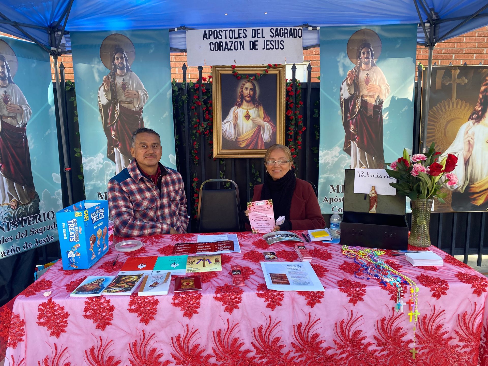 Two people seated at a memorial display with a portrait, candles, flowers, and blue draped banners.