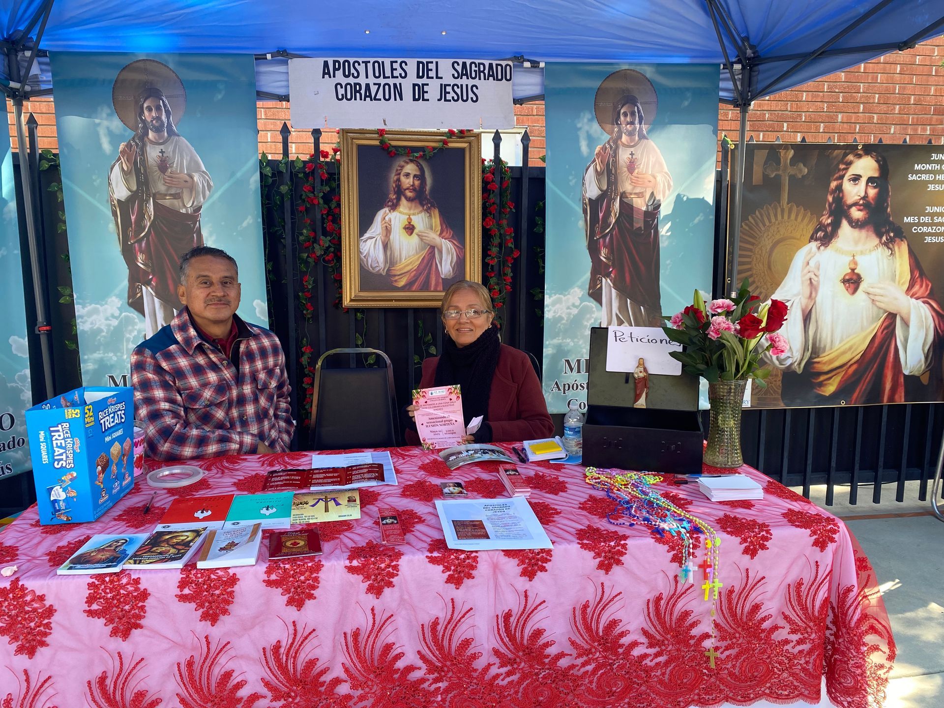 Two people seated at a red-draped table under a blue tent with religious posters and icons behind them