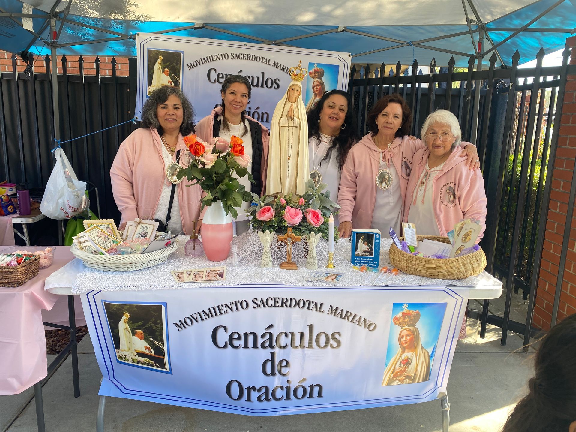 Five people at a Cénaculos de Oración booth with flowers, desserts, and a religious statue under a canopy