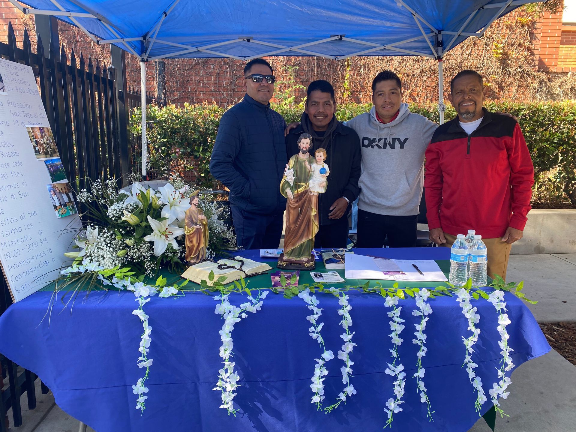 Four men behind a blue table display with a small religious statue and white floral decorations under a canopy.