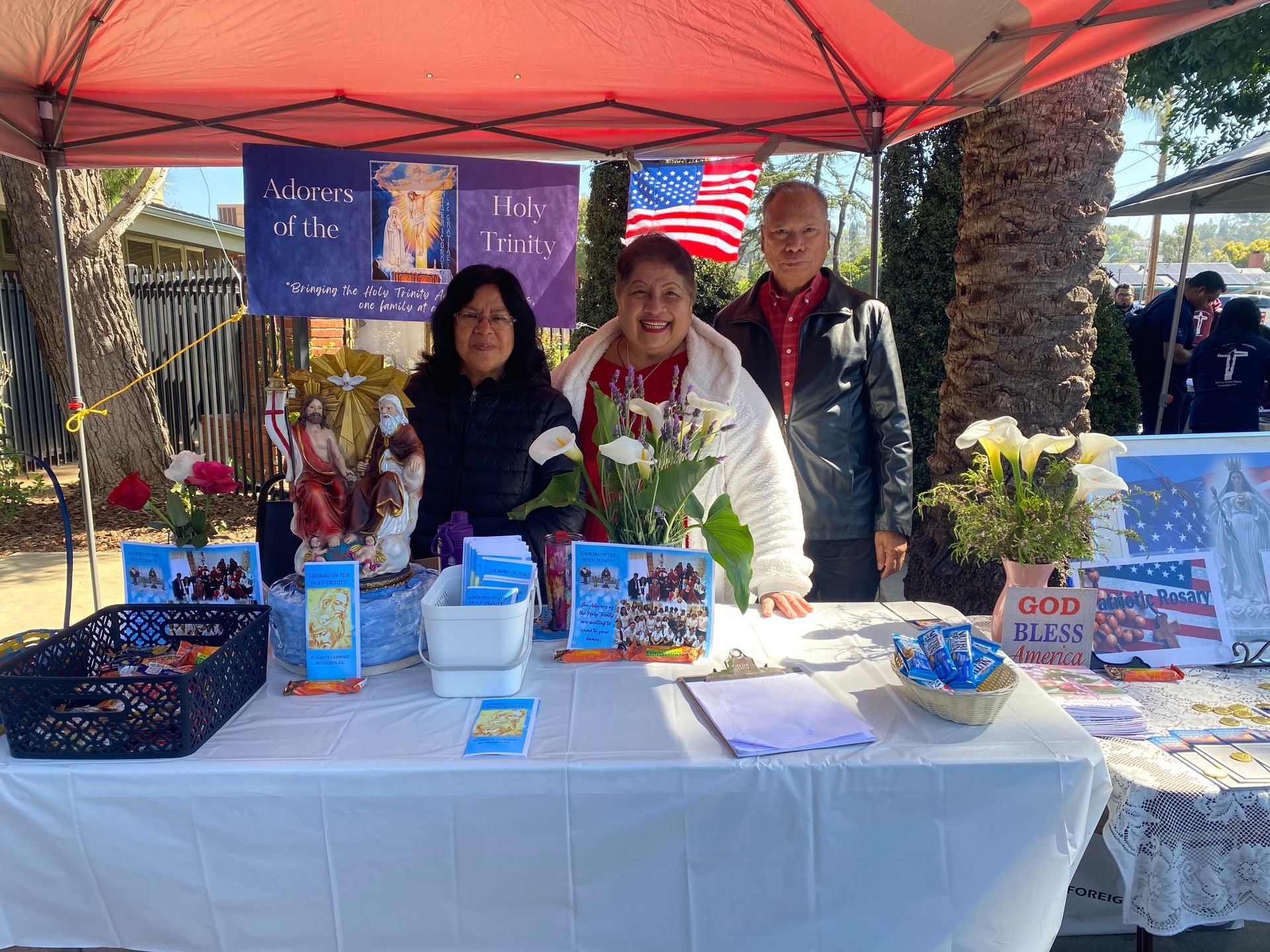 Three people at a table under a red canopy, with flyers, gifts, and a patriotic American flag backdrop.