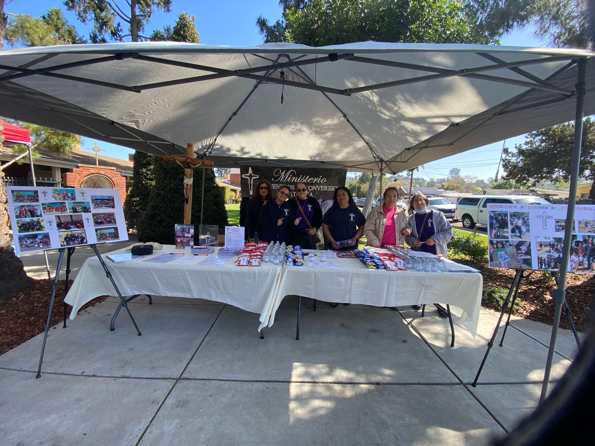 Outdoor community booth with volunteers behind a table under a white canopy, displaying brochures and items.