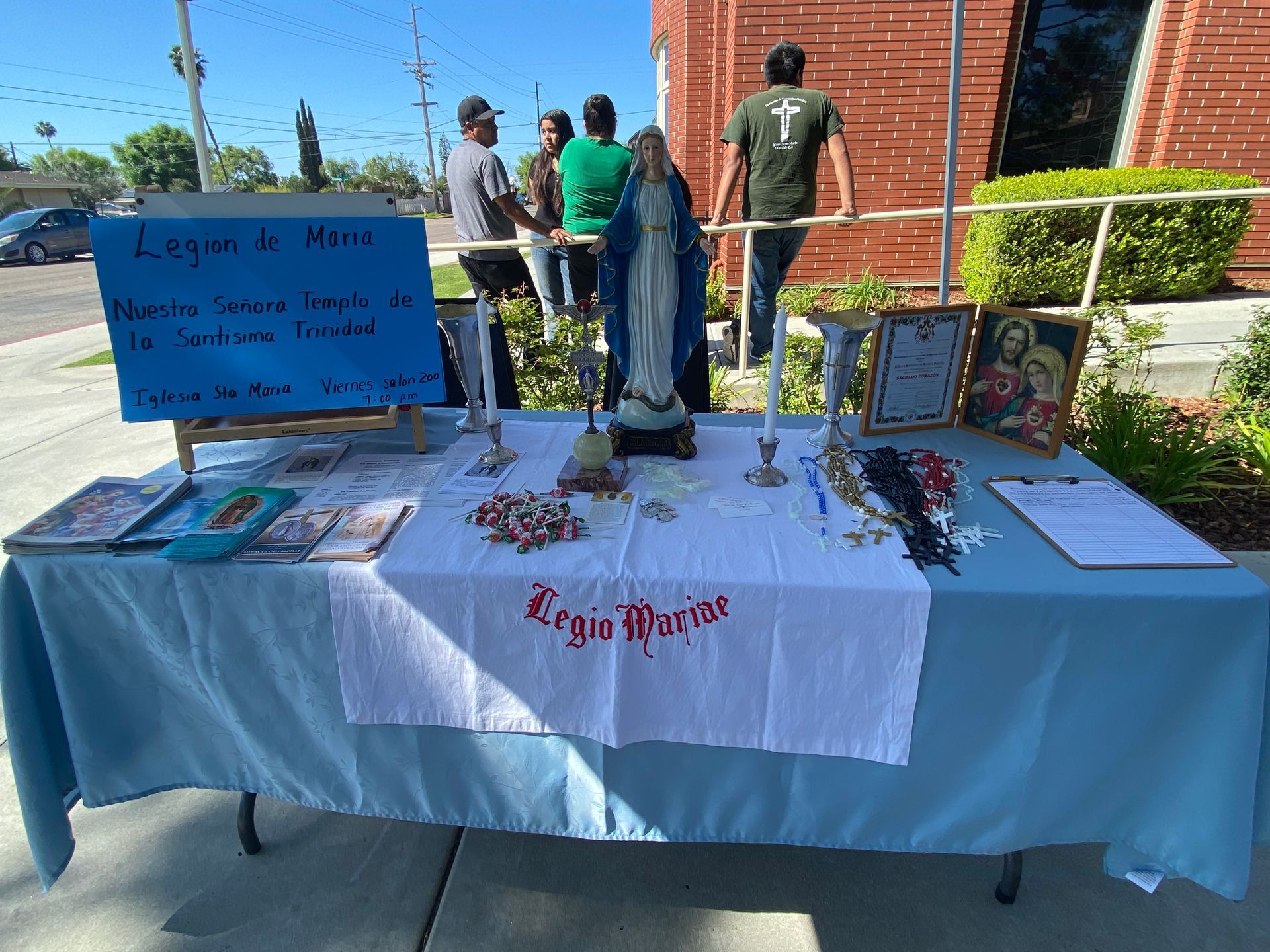 Outdoor memorial table with blue cloth, candles, flowers, framed photos, and a sign