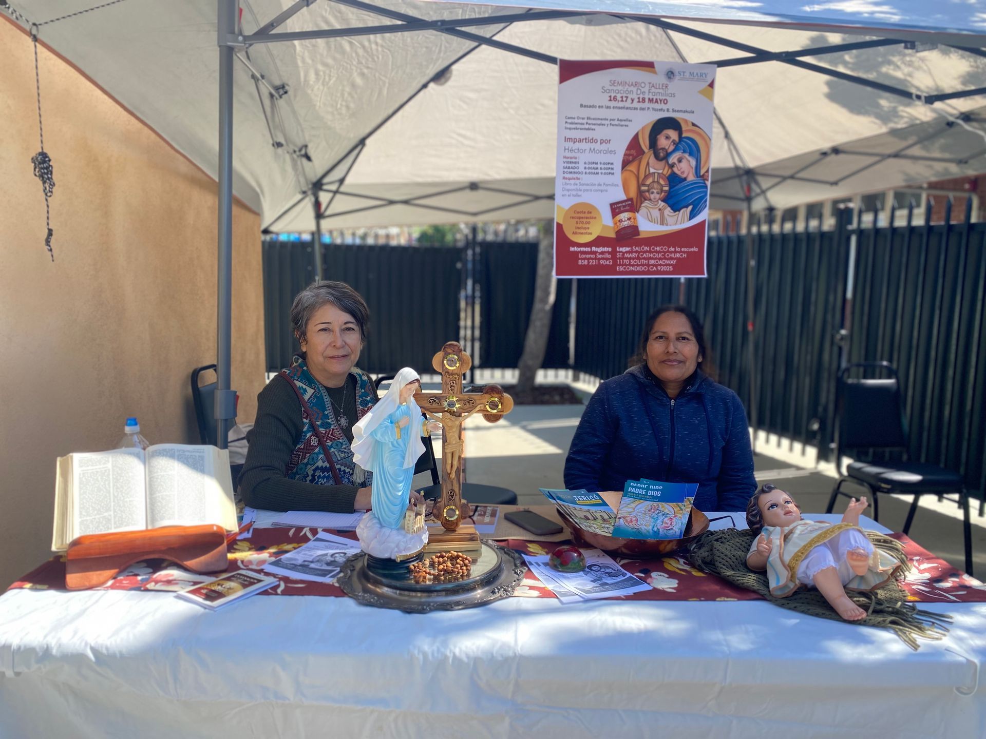 Two people seated at a market table under a canopy with snacks, books, and a religious statue.