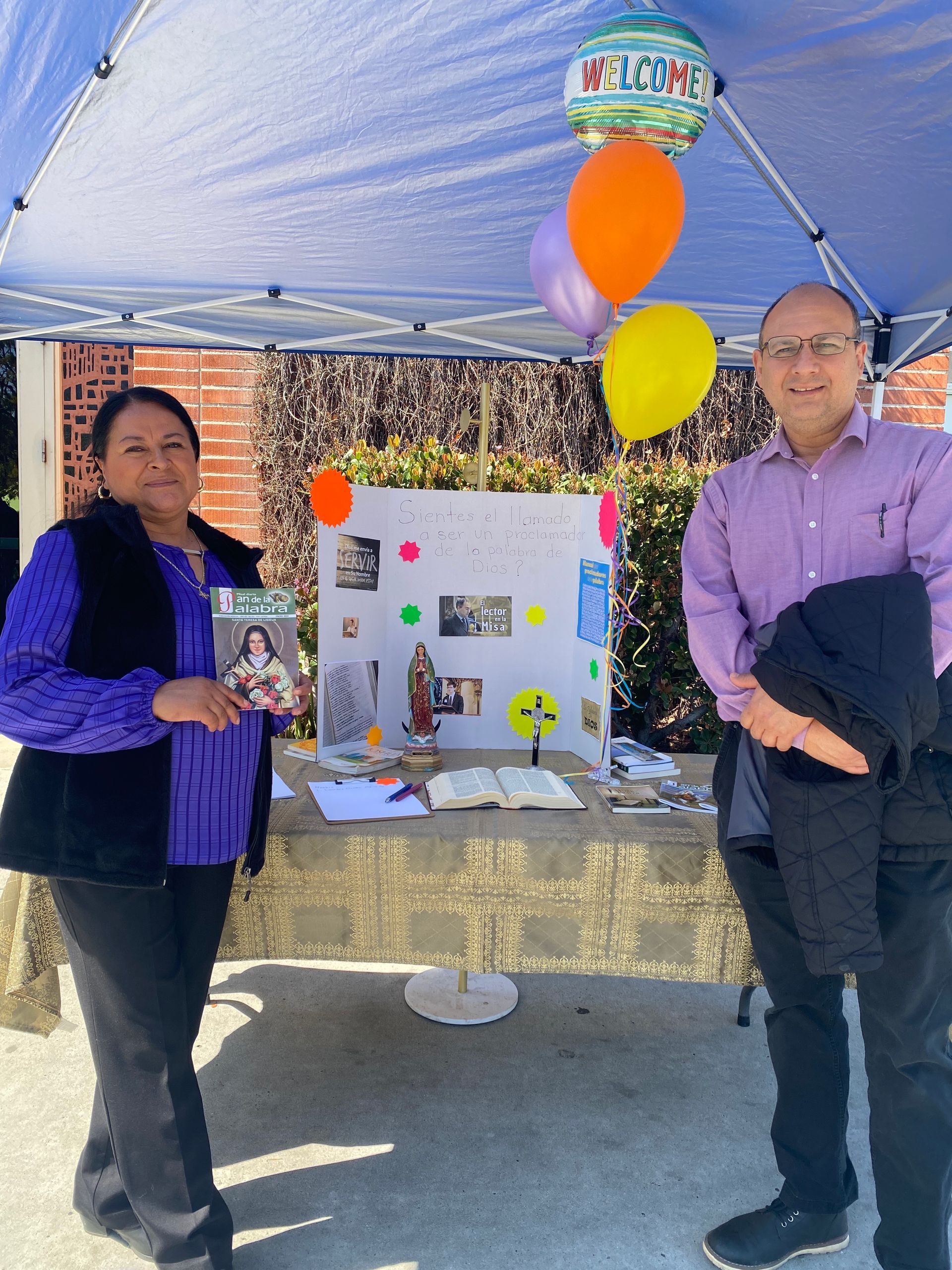 Two people standing beside a decorated display table under a white canopy with balloons and posters