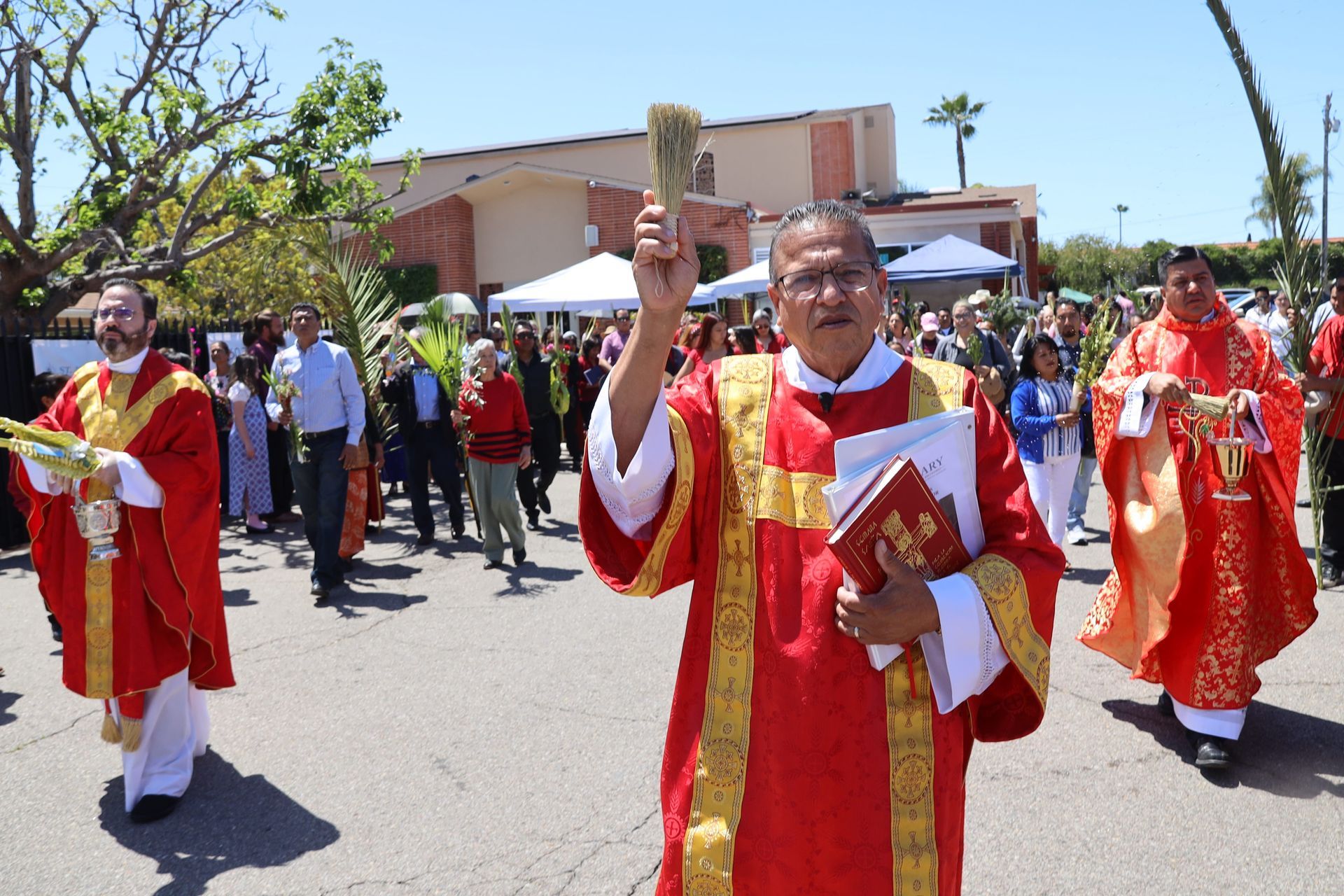 Clergy in red and gold vestments march in a church procession outdoors, one holding a book and blessing.