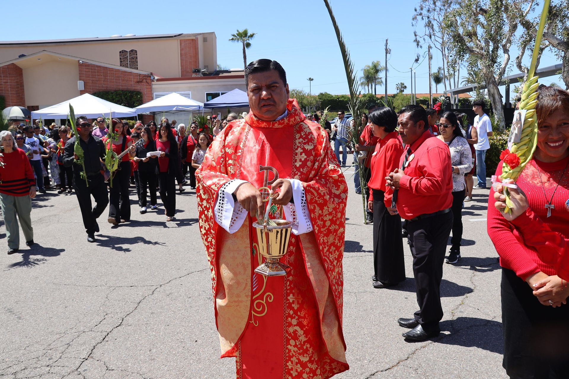 Outdoor procession with people in red traditional attire, led by a man holding ceremonial items