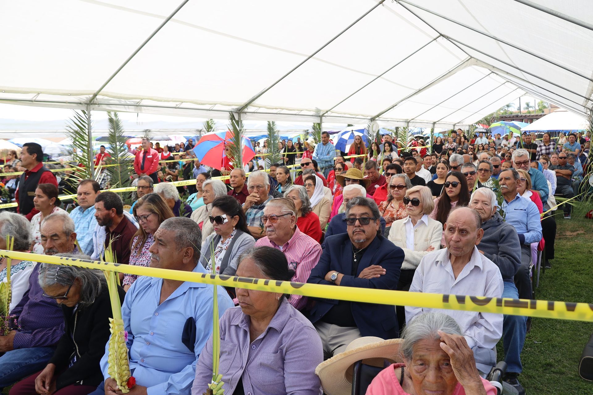Crowd seated under a white tent at an outdoor event, with yellow caution tape across the front row.