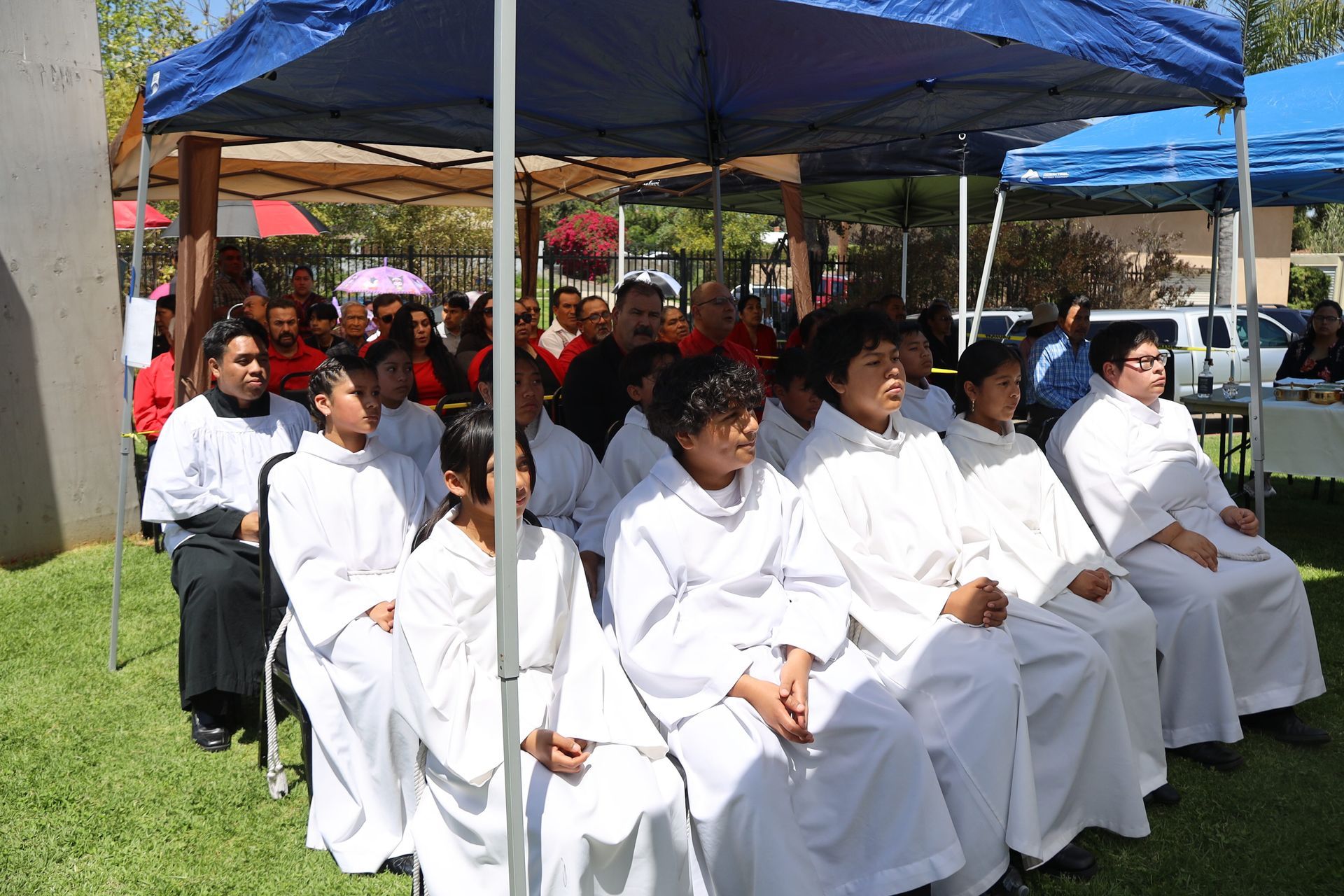 People seated outdoors under blue canopies at a formal gathering