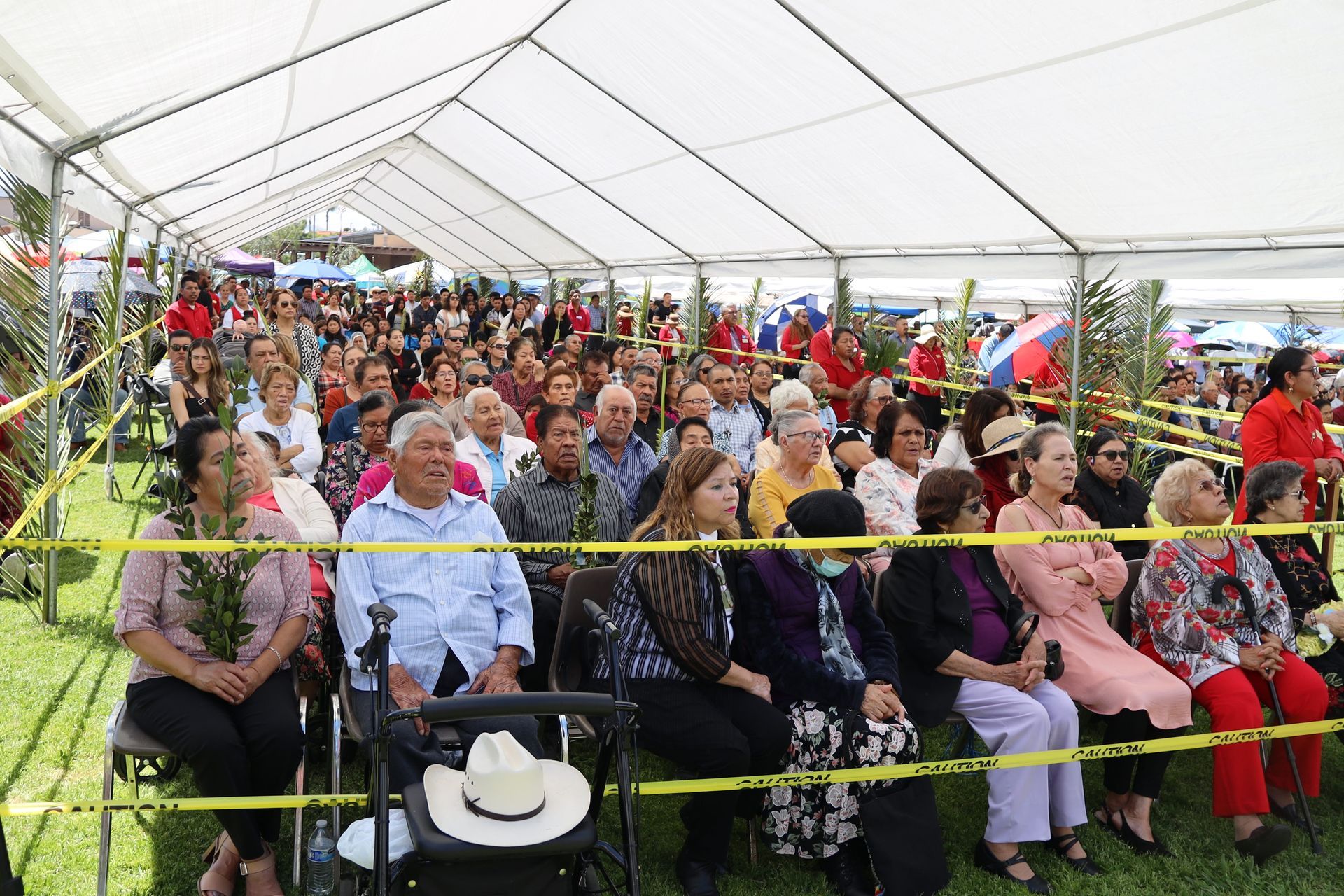Crowd seated under a large white tent at an outdoor event, with colorful clothing and red chairs