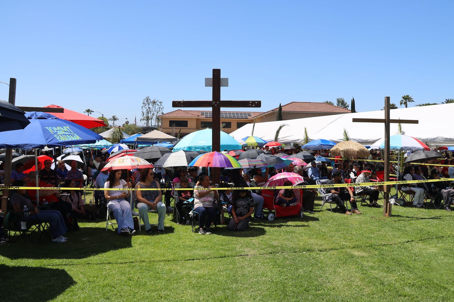 Crowd seated under colorful umbrellas on a grassy field with wooden crosses at an outdoor event