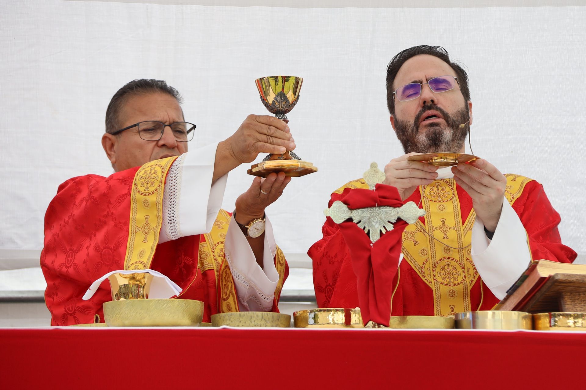 Two priests in red and gold vestments celebrate Mass, one lifting a chalice at the altar.