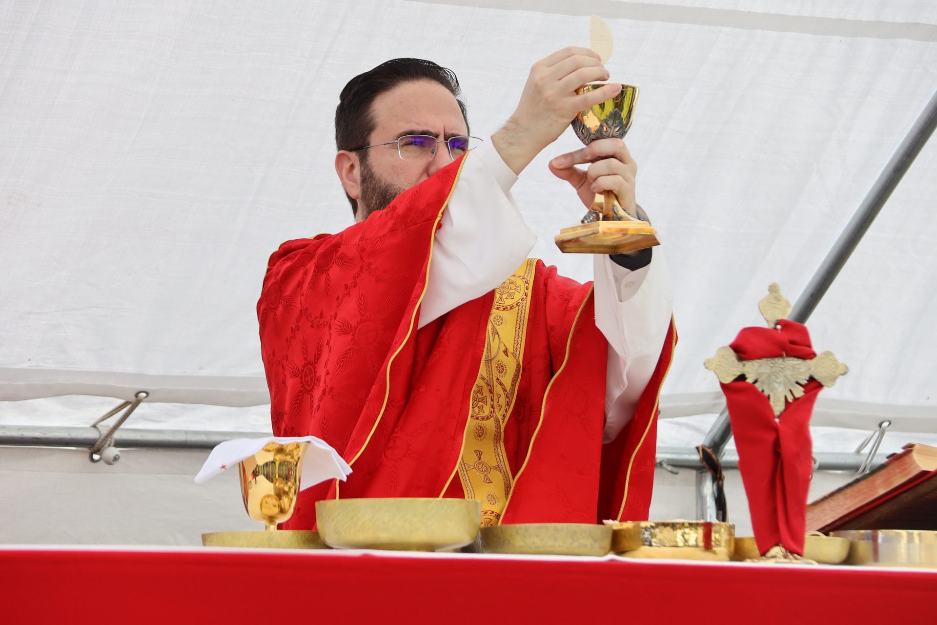 Priest in red vestments elevates a chalice during Mass under a white tent