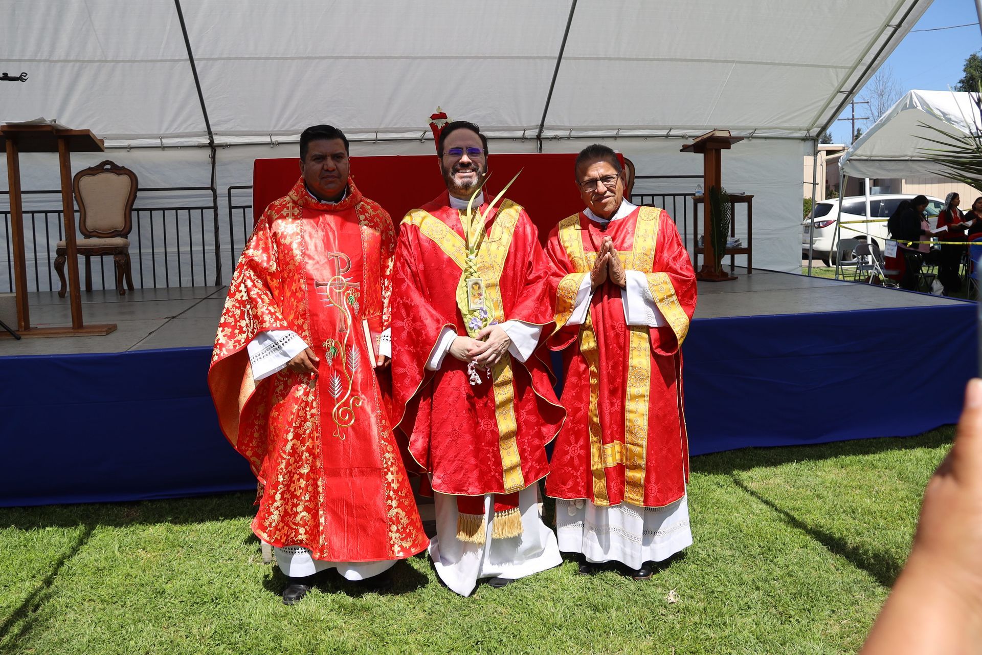 Three people in red and gold ceremonial robes standing on a stage outdoors under a white canopy.