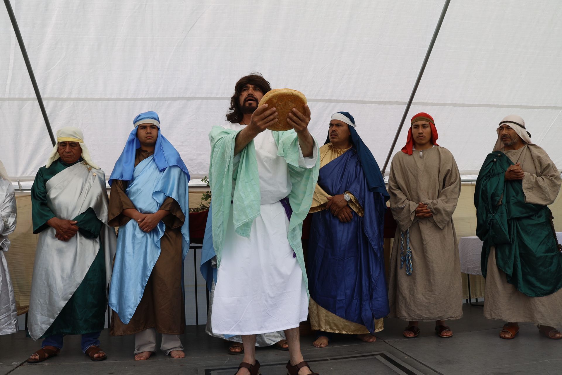 Seven people in desert robes and head coverings stand on a stage, one holding a loaf of bread.