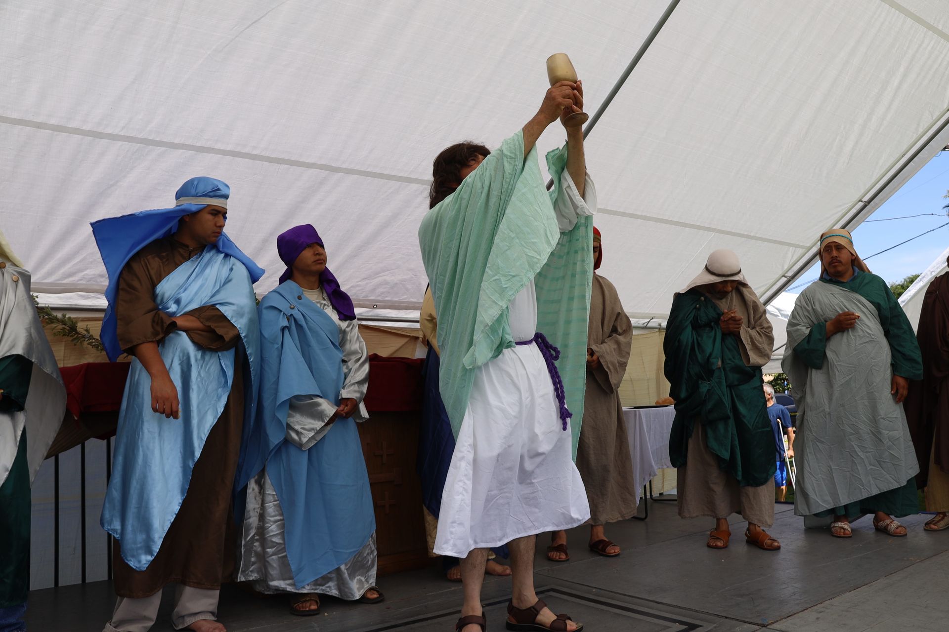 Performers in robes and headwraps dance on an outdoor stage under a white canopy