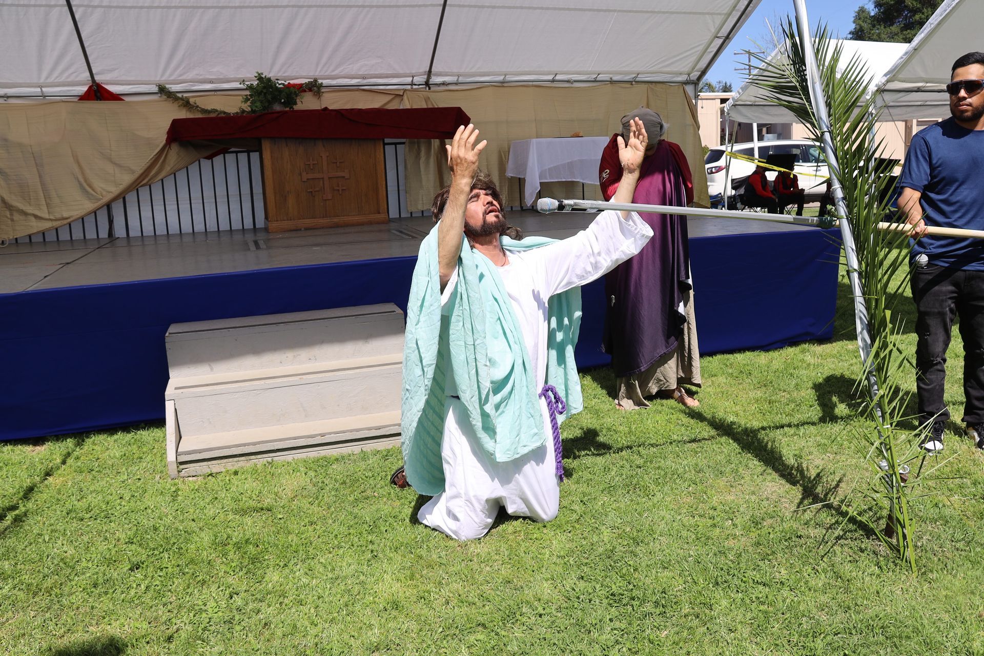Person kneeling with arms raised on a grassy stage under a tent, with others standing nearby