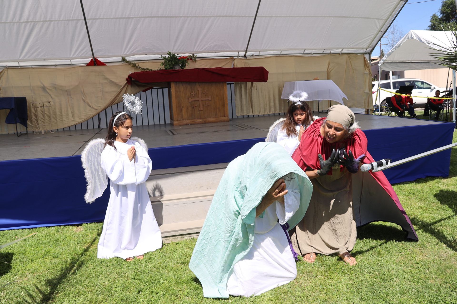 Children in angel costumes performing on a grassy outdoor stage under a white tent