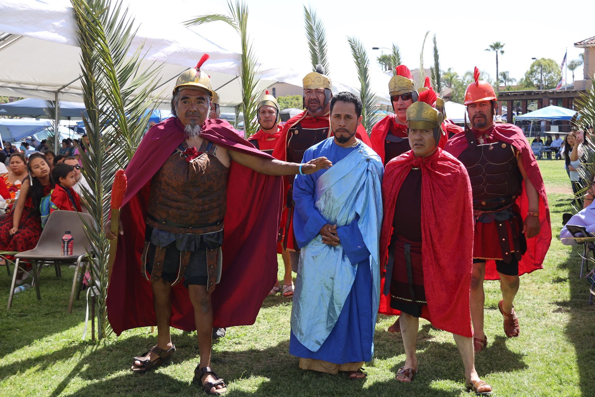 People in Roman-style costumes and red capes stand together on a grassy outdoor field.