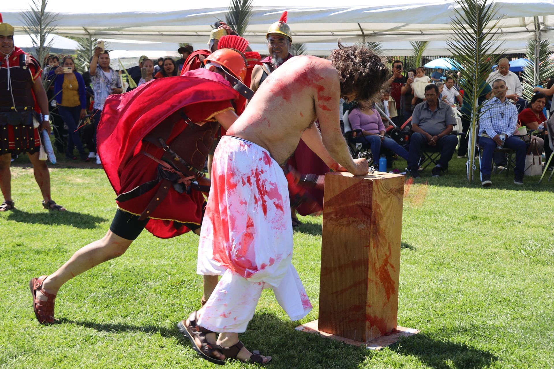 Two people in a reenactment scene, one leaning over a wooden block while the other supports him on a grassy field.
