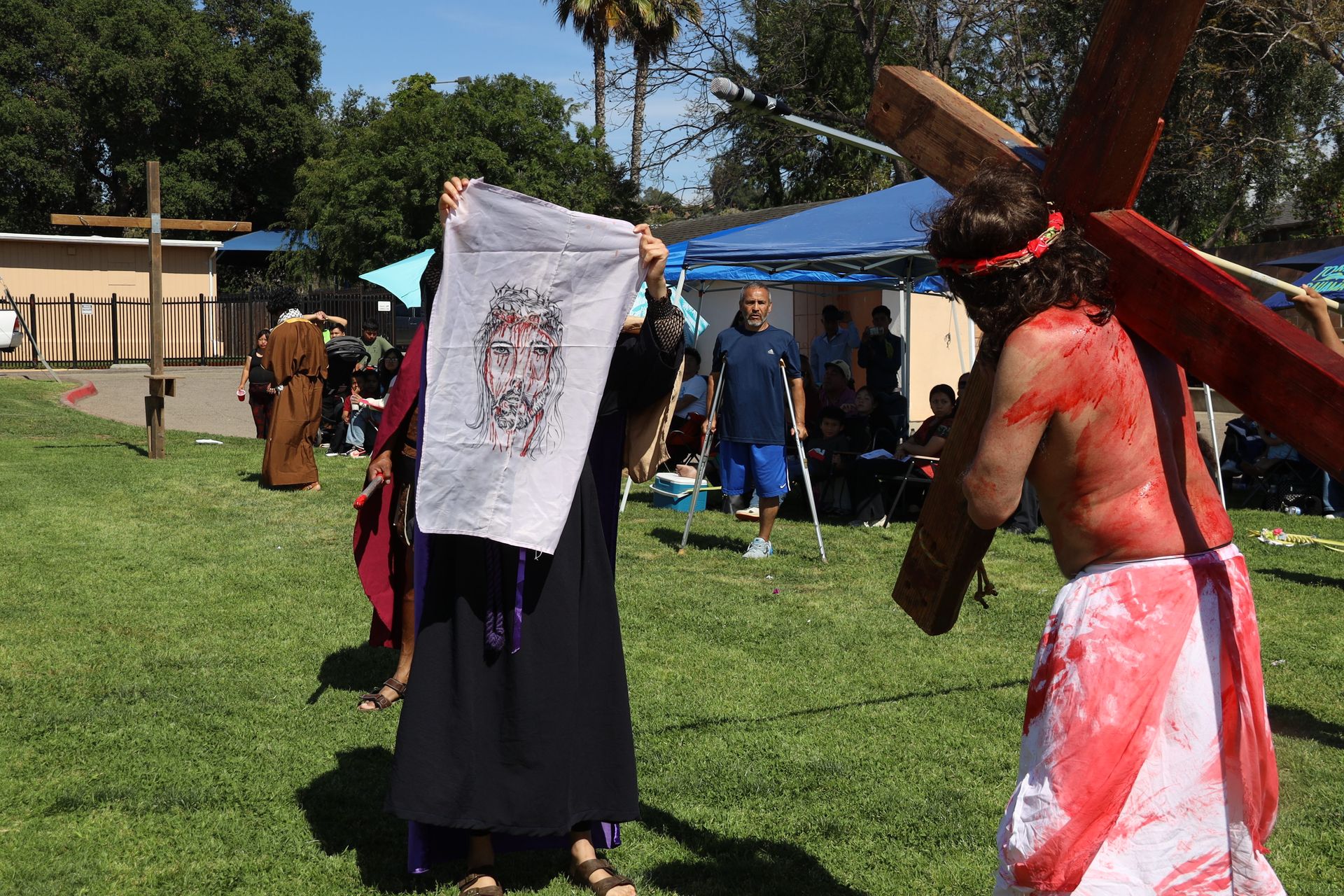 Costumed performers outdoors on grass, one holding a white banner and another carrying a wooden cross.