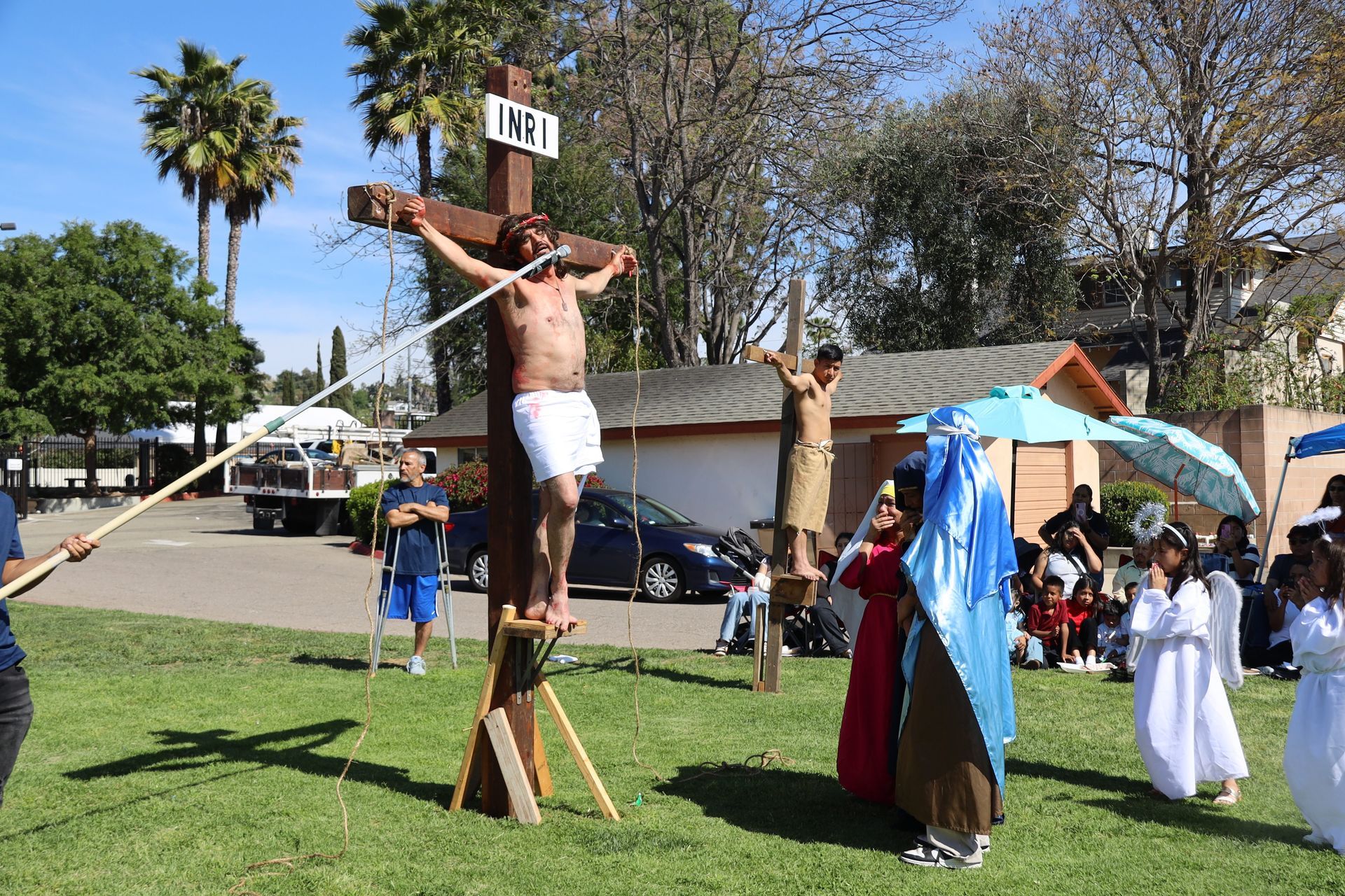 Outdoor reenactment with a crucifixion scene on a grassy field, with people gathered nearby