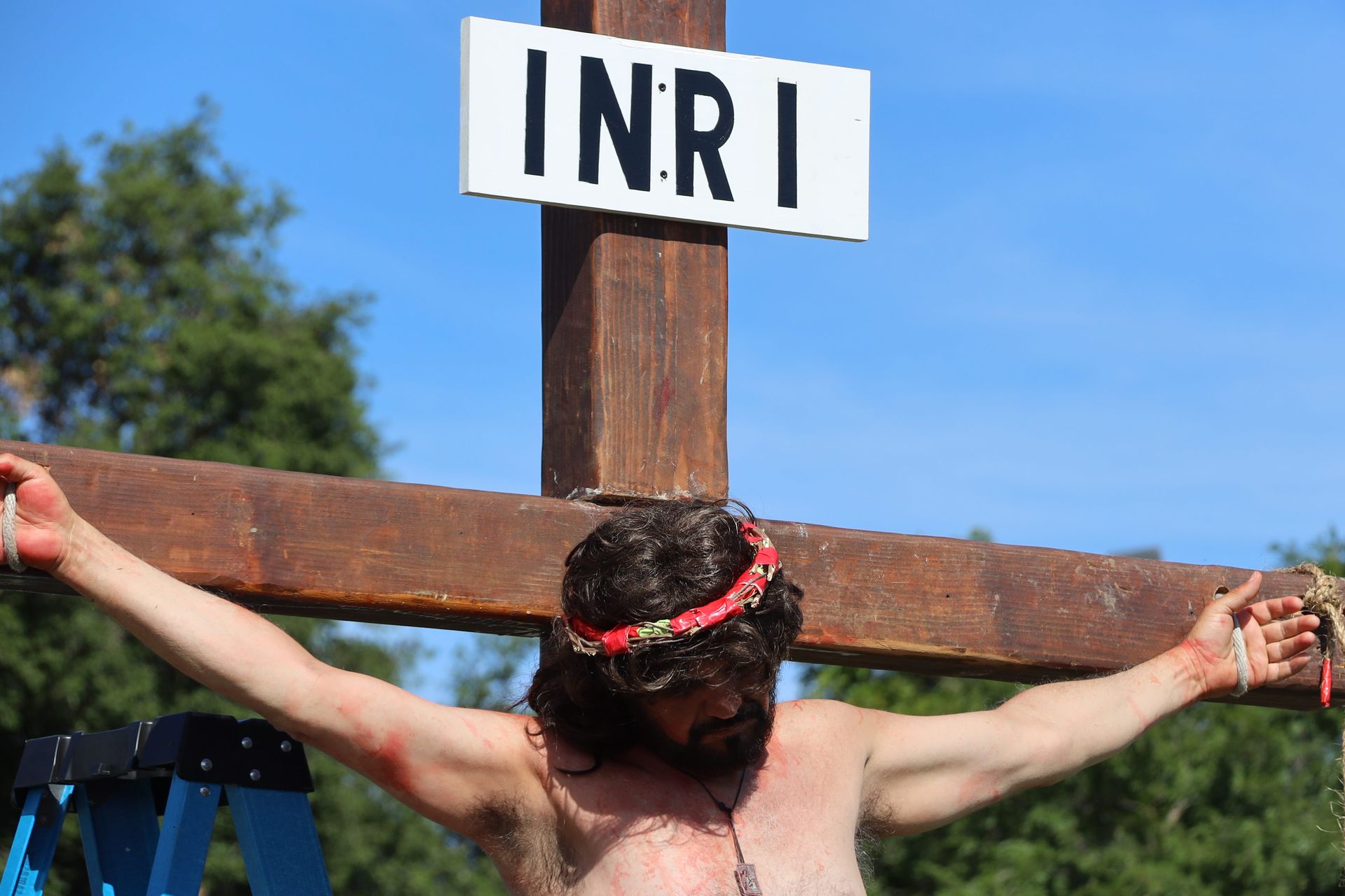 Man on a wooden cross with INRI sign, wearing a crown of thorns against a blue sky