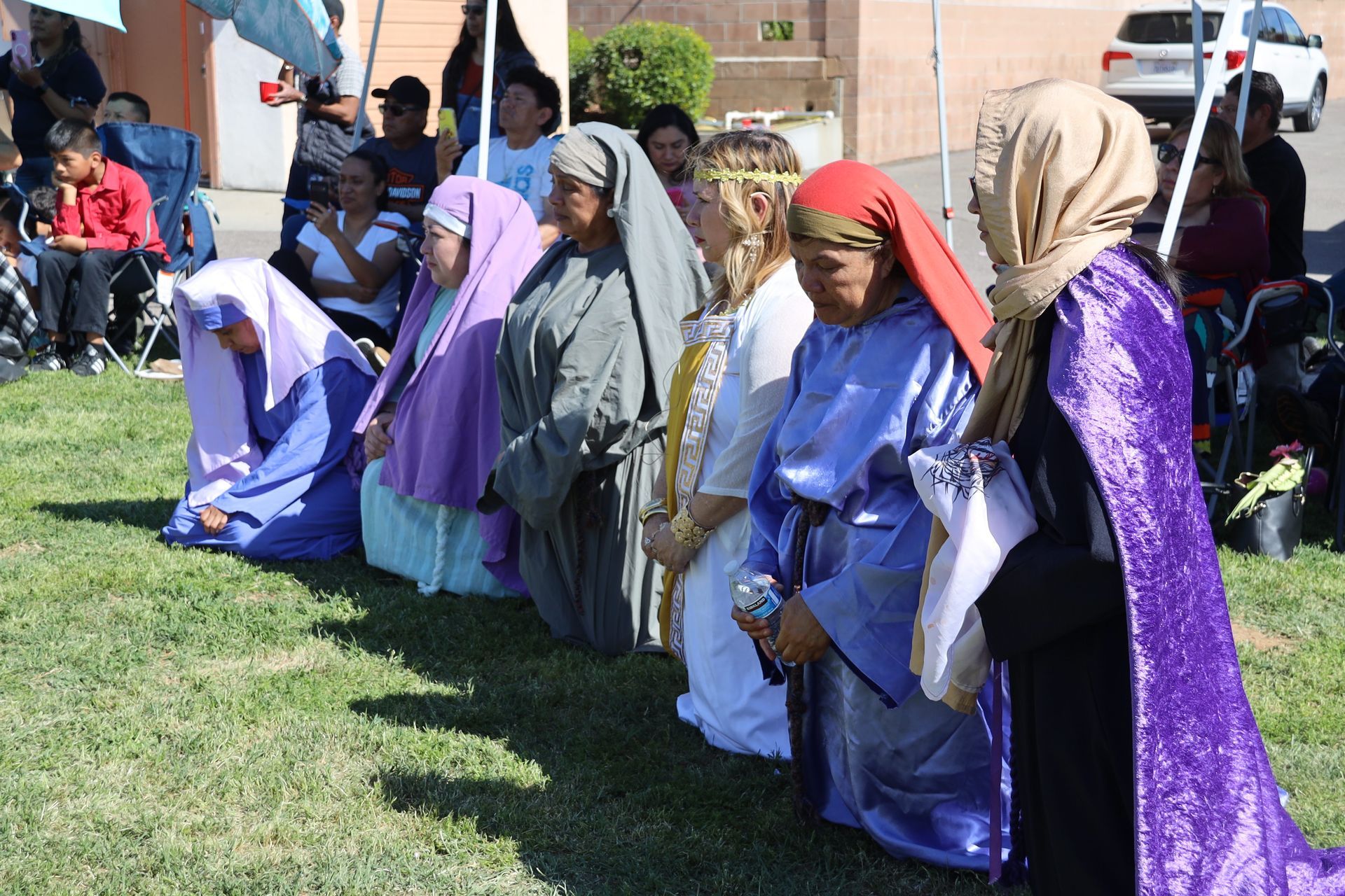 Children in colorful robes and headscarves kneel in a line on grass outdoors.