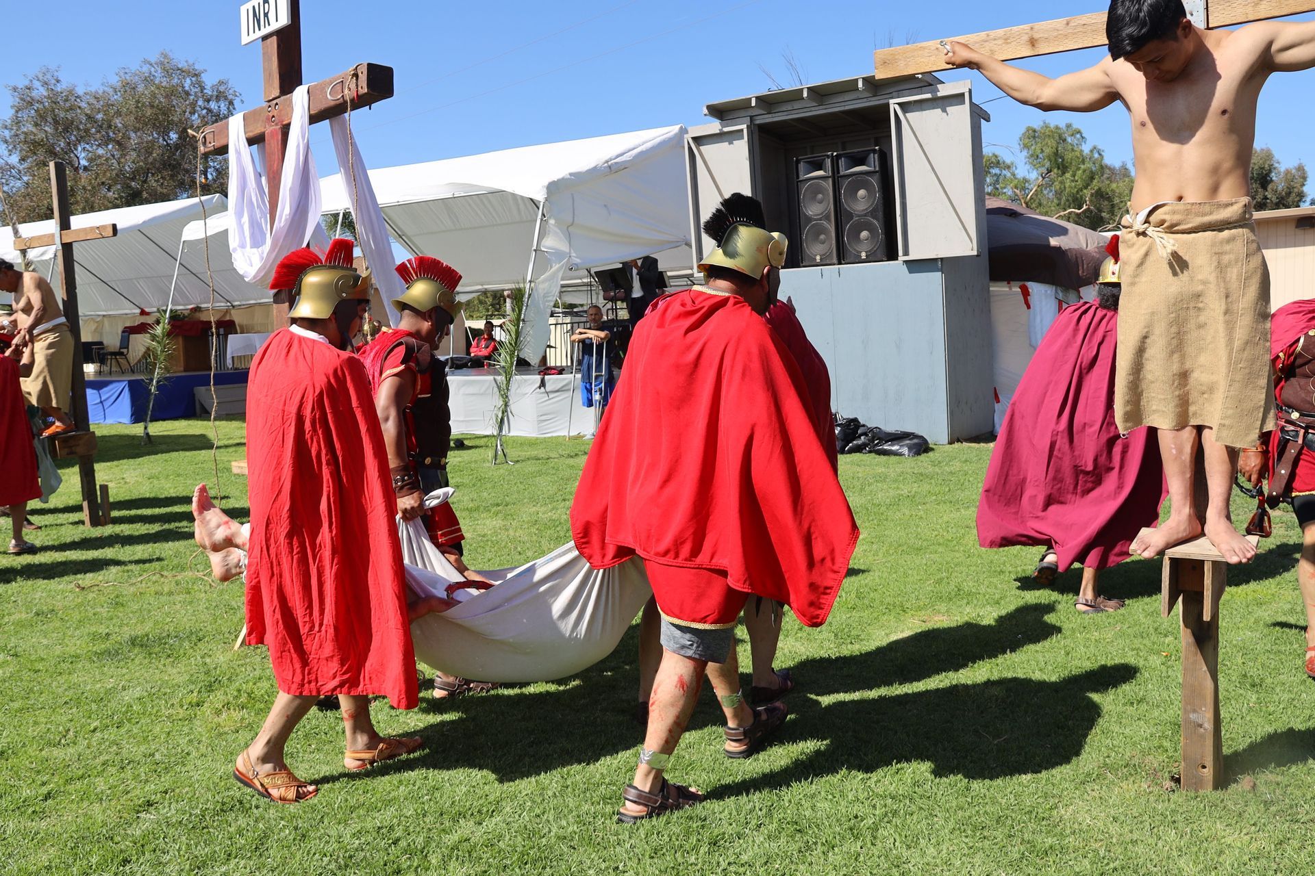People in red capes reenact a crucifixion scene outdoors on a grassy field.