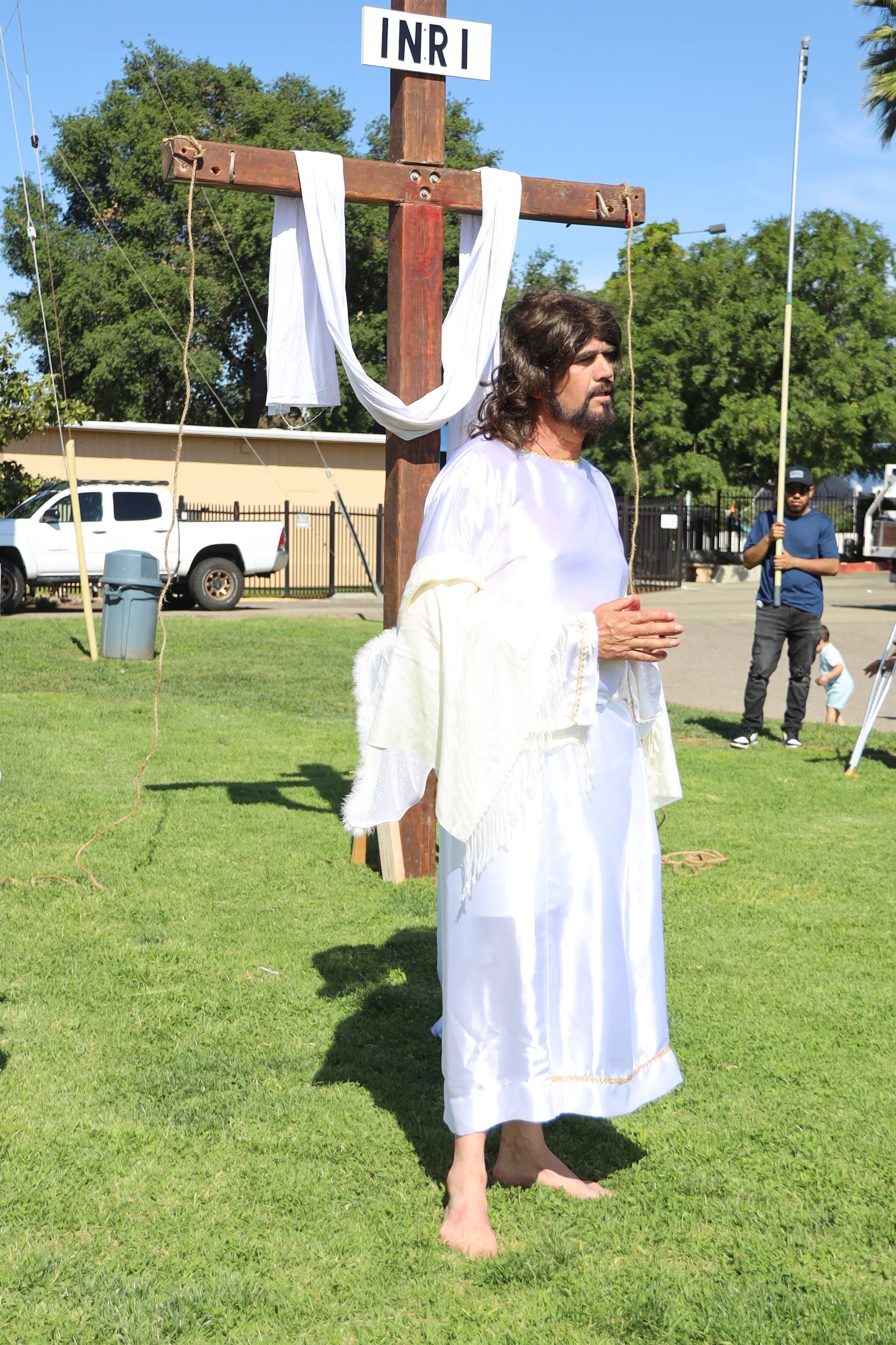 Person in white robe carrying a wooden cross outdoors on a grassy field during a religious event