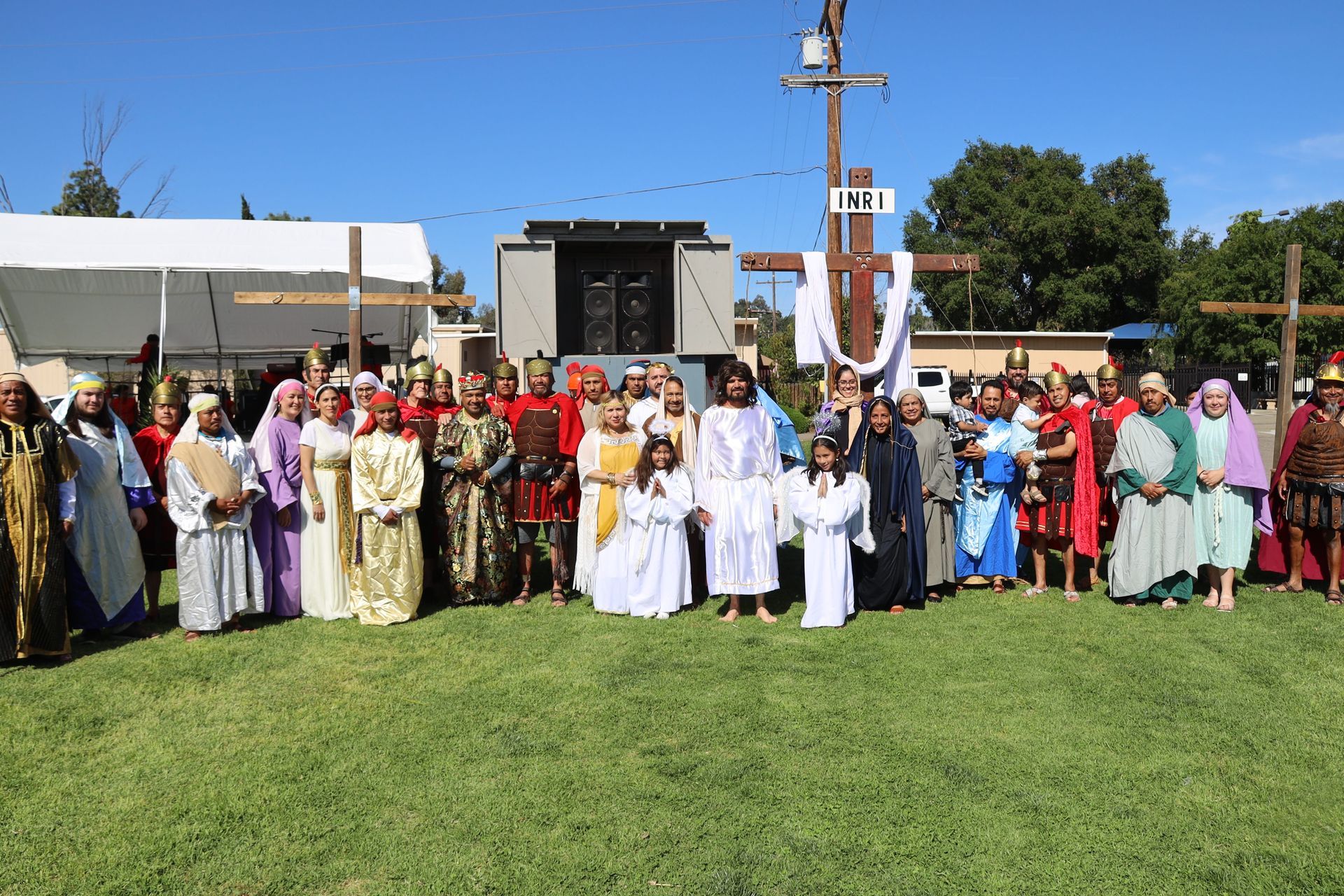 Large group standing in a grassy field outdoors near a stage, wearing colorful traditional clothing