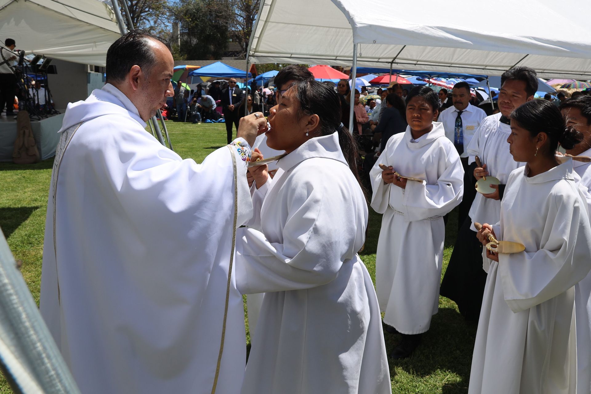 Clergy in white robes sharing communion outdoors under tents at a crowded gathering