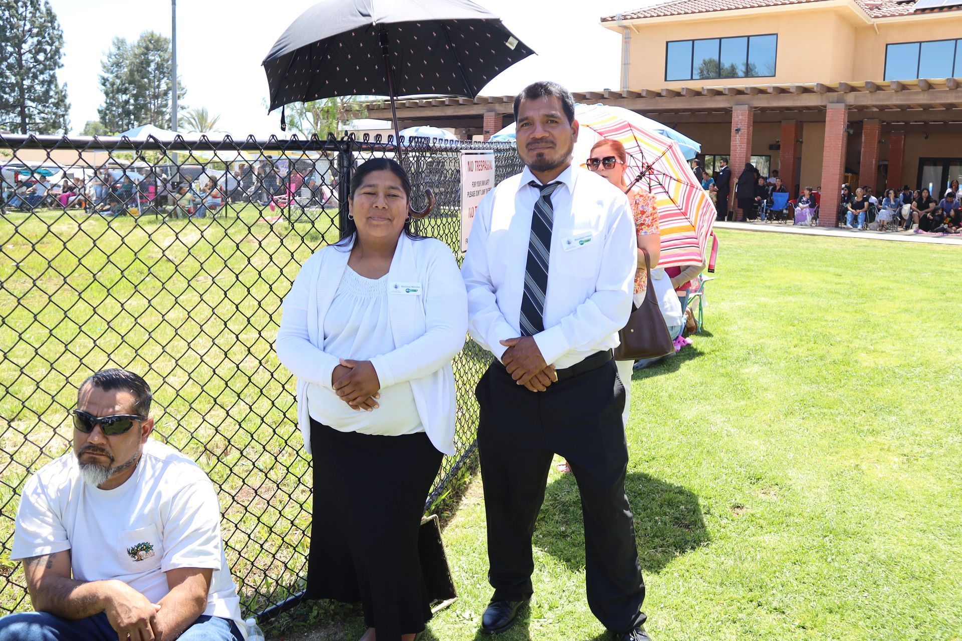 Two people standing by a fence outdoors, one holding an umbrella, with a building and crowd in the background.